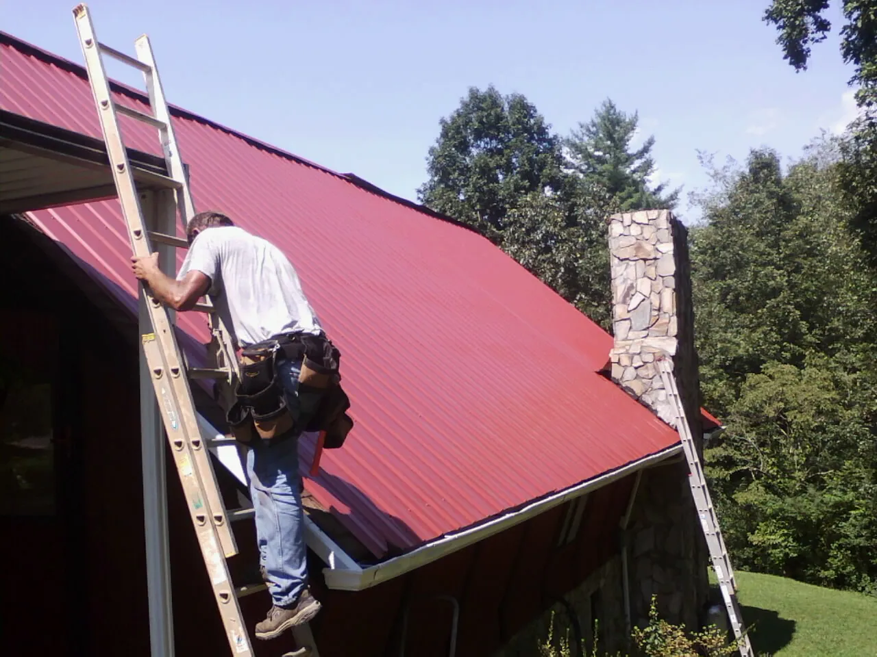 Roofer on a ladder beside a red metal roof and stone chimney — Soffit & Fascia Repair by Roofing Brothers in Shawsville